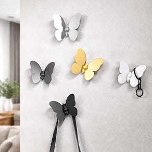 Pode incluir: Uma coleção de ganchos de parede em forma de borboleta em vários acabamentos metálicos: prata, dourado, preto e branco. Alguns ganchos seguram tiras pretas. Os ganchos são montados numa parede cinza claro, oferecendo uma solução de armazenamento decorativa e funcional.