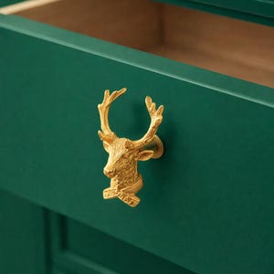 Puede incluir: Un tirador de cajón con forma de cabeza de ciervo dorada montado en un cajón verde oscuro. El cajón está abierto, revelando un interior marrón claro. La cabeza de ciervo tiene cuernos detallados y una superficie texturizada.