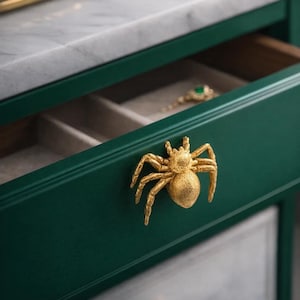 May include: A gold spider-shaped handle adorns a dark green drawer. The drawer is part of a cabinet with a white marble top. The drawer is open, revealing a jewelry tray. The spider handle provides a distinctive decorative touch to the cabinet.