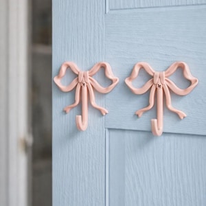 May include: Two pale pink bow-shaped hooks fixed to a light blue wooden door. The hooks are in the shape of a bow with a curved hook. The door has a vertical wood grain texture. The hooks are likely for hanging items.
