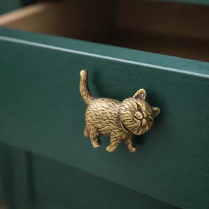 May include: A brass-colored cat-shaped drawer pull is attached to a dark green drawer. The cat is in a walking pose with its tail up and its eyes closed. The drawer is open, revealing the interior.