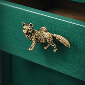 Könnte beinhalten: Antiker, messingfarbener Schubladengriff in Fuchsform, montiert auf einer dunkelgrünen Schublade. Der Fuchs ist in einer Laufpose dargestellt, mit detaillierten Fellstrukturen. Ein passender Messingknopf ist unten sichtbar.