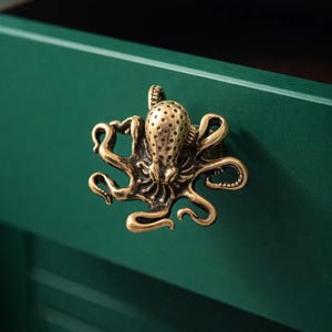 May include: A brass octopus cabinet knob with a detailed design. The knob is mounted on a dark green cabinet door. The octopus has a textured head and curling tentacles, adding a unique nautical touch to the furniture.