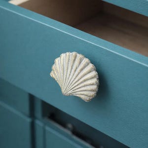 Pode incluir: Puxador de armário em forma de concha com acabamento texturizado branco sujo e detalhes dourados. O puxador está montado numa porta de armário azul-petróleo, com uma gaveta parcialmente aberta. Ferragens decorativas.