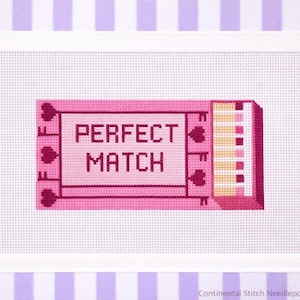 Puede incluir: Un lienzo de punto de aguja rosa con las palabras "PERFECT MATCH" en un diseño rectangular. El diseño incluye formas de corazón y una ilustración de cerilla. El lienzo está enmarcado en blanco y sobre un fondo a rayas.
