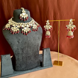 May include: A gold-coloured necklace, earrings, and headpiece set. The jewellery features clear, teardrop-shaped stones and small red beads. The necklace and headpiece are displayed on a grey bust, while the earrings are on a gold stand.