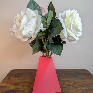 May include: A bright pink geometric vase displays white roses and green foliage. The vase features a modern, angular design. The flowers are full and lush, contrasting with the vase's bold colour and shape. The arrangement is on a wooden surface.