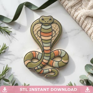 May include: A cobra-shaped biscuit decorated with royal icing in shades of olive green, muted orange, and cream. The design features a detailed hood, a coiled body, and dark grey outlines that define the snake's scales and facial features.