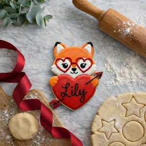 Puede incluir: Una galleta decorada con forma de zorro con gafas de corazón y un corazón rojo con el nombre "Lily". Una flecha atraviesa el corazón. Otros elementos de repostería incluyen un rodillo, masa y una cinta roja.