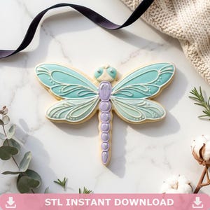 May include: A dragonfly-shaped cookie with teal and lavender icing. The wings are a gradient of teal and light blue, with white decorative lines. The body is lavender, and the cookie sits on a white marble surface. Text at the bottom reads "STL INSTANT DOWNLOAD".
