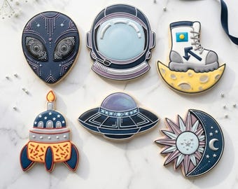 May include: Assortment of space-themed cookies, including an alien, astronaut helmet, space boot on a crescent moon, rocket ship, UFO, and a sun-moon design. The cookies are decorated with blue, white, yellow, and orange icing.