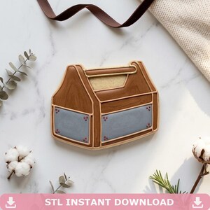 May include: A cookie shaped like a brown toolbox with a handle and grey panels featuring small red dots at the corners. The cookie has a wood grain texture and is outlined in light tan icing. The text STL INSTANT DOWNLOAD is at the bottom.