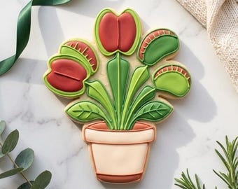 May include: A decorated sugar cookie shaped like a potted Venus flytrap plant. The cookie features a light beige pot, vibrant green stems and leaves, and several trap heads in shades of deep red and bright green with visible serrated edges.