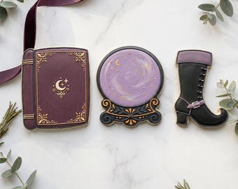 May include: Cookies in the shape of a book, crystal ball, and boot. The book is dark purple with gold accents and a crescent moon design. The crystal ball is purple with gold swirls and sits on a black and gold stand. The black boot has a purple cuff.