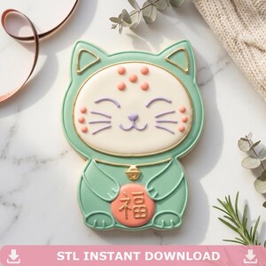 May include: A cat-shaped cookie decorated with mint green icing, a cream-colored face, and a gold-outlined bell. The cat holds a round peach-colored coin featuring the gold Chinese character for fortune, with small peach dots on its forehead and cheeks.