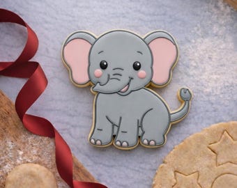 May include: A gray elephant-shaped cookie with pink ears and a smiling face. The cookie is decorated with black icing details and sits on a light gray surface. A wooden rolling pin, star-shaped cookie dough, and a red ribbon are also present.