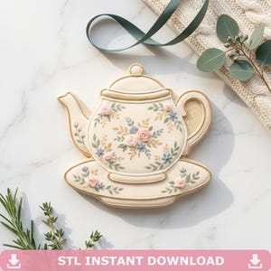 May include: A cream-colored teapot-shaped cookie with a gold outline, decorated with pink and blue floral designs. The cookie sits on a matching saucer. A green ribbon and sprigs of greenery are in the background.