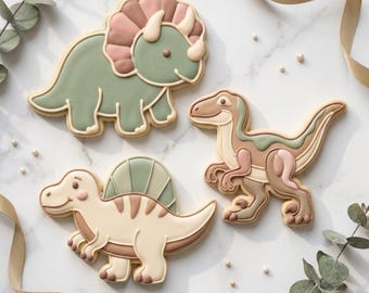 May include: Three dinosaur-shaped cookies. A green triceratops with a pink frill, a tan and green velociraptor, and a tan and brown spinosaurus. The cookies are outlined in a light brown color. Text at the bottom reads "STL INSTANT DOWNLOAD".