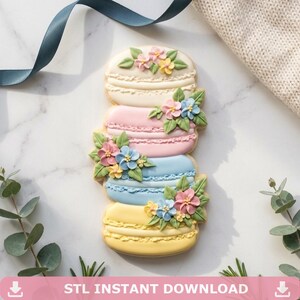 Puede incluir: Una pila vertical de cuatro galletas decoradas con forma de macarons en colores crema, rosa, azul y amarillo. Cada galleta está adornada con pequeñas flores de glaseado de colores en tonos rosa, amarillo y azul, junto con hojas de glaseado verde.