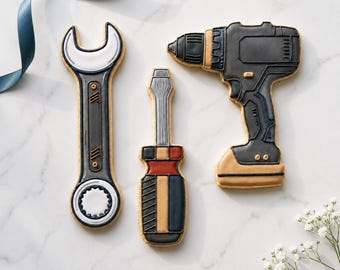 May include: Three tool-shaped cookies: a wrench, a screwdriver, and a drill. The wrench is gray and white, the screwdriver has a red, black, and tan handle, and the drill is gray and tan. The cookies are arranged on a white marble surface.