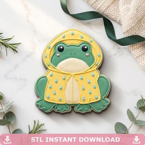 May include: A frog-shaped cookie decorated with royal icing, featuring a green frog wearing a yellow raincoat with blue raindrop patterns. The frog has a cream-colored belly, black eyes, and pink cheeks. The design is centered and flat.