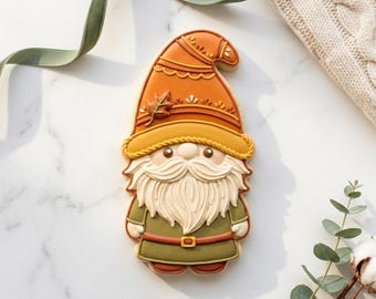 May include: A gnome-shaped cookie decorated with royal icing. The gnome wears a tall orange hat with a yellow brim and a small leaf detail, a long cream-colored beard, and a dark olive green tunic with a brown belt and matching boots.
