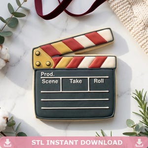 May include: A cookie shaped like a film clapperboard, with a dark green base and a striped top in red, yellow, and white. The clapperboard has white lines for writing and the words "Prod., Scene, Take, Roll".