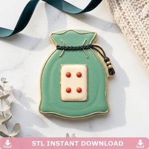 May include: A green biscuit shaped like a drawstring pouch with a gold-outlined cream-coloured rectangle in the centre featuring four red dots. The pouch has a black braided drawstring with a tassel and gold beads, all finished with gold piping.
