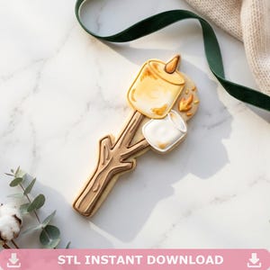 May include: A decorated sugar cookie shaped like a wooden stick with two marshmallows attached. One marshmallow is toasted golden brown with a small flame detail, while the other is white with brown accents. The cookie features detailed icing.