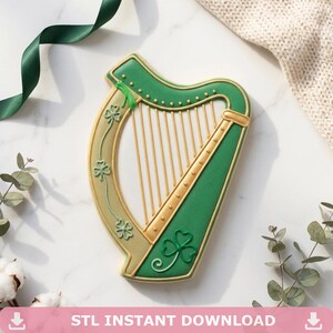 May include: A harp-shaped cookie decorated with green and gold icing. The frame features raised shamrock details in green, while the strings are represented by thin gold lines. The design includes a gold border and a green body section.