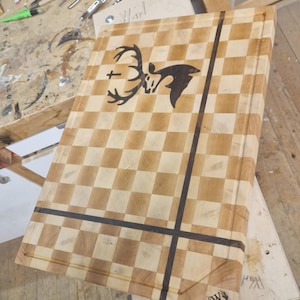 Puede incluir: Tabla de cortar de madera tipo « butcher block » con un diseño a cuadros y una silueta de ciervo. La tabla tiene líneas oscuras y un logotipo en la esquina. Se ven herramientas de tallado en el fondo.