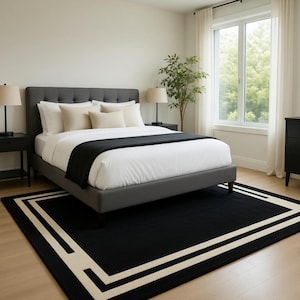 May include: A modern bedroom featuring a gray upholstered bed with white bedding and a black throw. Two black nightstands with lamps flank the bed. A black rug with a cream border sits on the hardwood floor.
