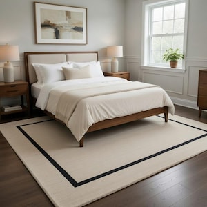 May include: A bedroom scene featuring a bed with white bedding, a wooden frame, and a beige headboard. A beige rug with a black border is on the floor. Two bedside tables with lamps flank the bed. A painting hangs above the bed.