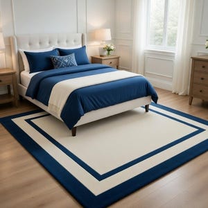 May include: A bedroom scene featuring a bed with navy blue bedding, pillows, and a decorative pillow. A cream-colored throw blanket is draped across the bed. A large rectangular rug with a cream center and navy blue border is on the floor.