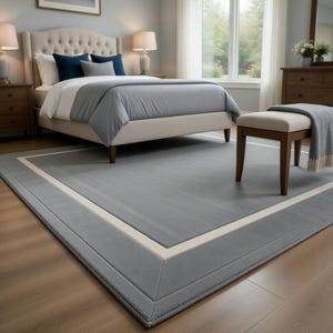May include: A gray rectangular area rug with a cream-colored border in a bedroom setting. The rug is placed on a hardwood floor, and a bed with gray bedding is visible in the background. The room is well-lit with natural light.