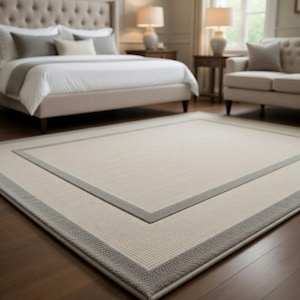 May include: A rectangular area rug with a neutral color palette. The rug features a light beige center with a gray border. The rug is placed on a hardwood floor in a bedroom setting, with a bed and sofa visible in the background.