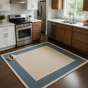 May include: A rectangular area rug with a cream-colored center and a wide, blue border. The rug is placed on a dark wood floor in a kitchen setting. Two pairs of beige slippers are on the rug.