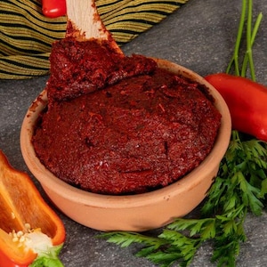 Op de afbeelding: Een terracotta kom gevuld met een levendige rode pasta, waarschijnlijk een saus op basis van tomaat of paprika. Een houten lepel rust in de kom, met een deel van de pasta erop. Rode paprika's, peterselie en een gesneden oranje paprika zijn rond de kom gerangschikt.