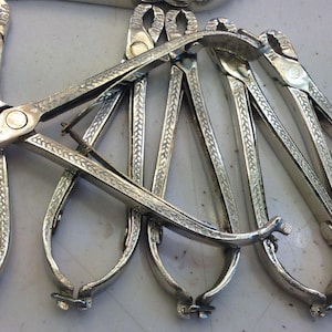 May include: A collection of silver-colored metal tools, possibly pliers or tongs, with intricate decorative patterns. The tools have a hinged design with curved handles and a central pivot point. The word "HAVE" is stamped on the tools.