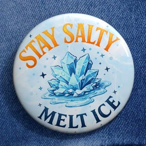 Może przedstawiać: Okrągła, biała przypinka z napisem "Stay Salty Melt Ice" w kolorze pomarańczowym i niebieskim. Przypinka przedstawia niebieski kryształ z akcentami wody i gwiazd. Przypinka jest przypięta do dżinsowej kurtki.