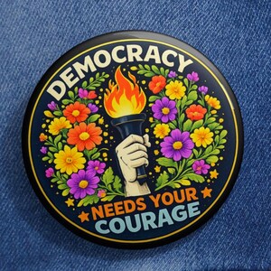May include: A round, black button with the words "DEMOCRACY" and "NEEDS YOUR COURAGE" encircling a torch held by a hand, surrounded by colorful flowers. The button is pinned to a blue denim jacket.