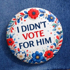 May include: A circular badge with the words "I DIDN'T VOTE FOR HIM" in blue and red, surrounded by a floral wreath of red, white, and blue flowers. The badge is attached to a denim jacket.