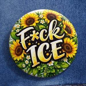May include: A round button with a sunflower design and the words "F*ck ICE" in a bold, stylized font. The button is surrounded by yellow sunflowers, white daisies, and green leaves, set against a denim jacket.