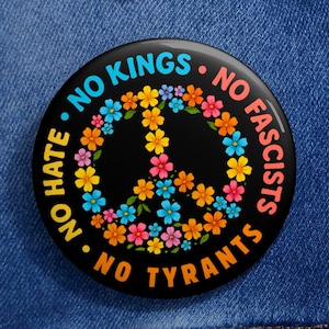 May include: A round black button with a peace sign made of colorful flowers. The button has the words "NO HATE", "NO KINGS", "NO FASCISTS", and "NO TYRANTS" in a circular pattern around the peace sign.