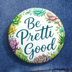 Puede incluir: Un botón redondo con el texto "Be Pretti Good" en escritura verde azulado, rodeado por una corona de suculentas coloridas. El botón está sobre una chaqueta vaquera.