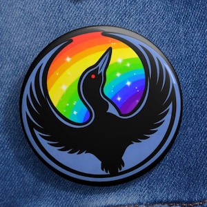 Puede incluir: Un botón redondo con un diseño de pájaro negro sobre un fondo de arcoíris. El pájaro tiene las alas extendidas y un ojo rojo. El pin está sobre una chaqueta vaquera azul.