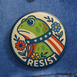 May include: A round button featuring a cartoon frog with a patriotic design. The frog is green with red spots and wears a scarf with stars and stripes. The word "RESIST" is printed at the bottom, surrounded by flowers.