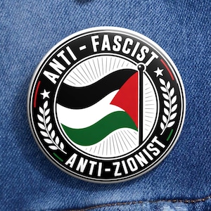 Puede incluir: Un botón redondo con las palabras "ANTI-FASCIST" y "ANTI-ZIONIST" alrededor de un diseño de bandera. La bandera tiene los colores negro, blanco, rojo y verde. El botón está fijado a una chaqueta vaquera azul.