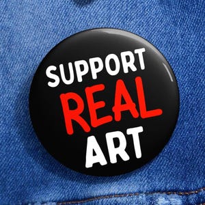Könnte beinhalten: Ein runder, schwarzer Button mit den Worten "SUPPORT REAL ART" in weißer und roter Schrift. Der Button ist an einer blauen Jeansjacke befestigt.
