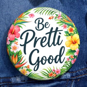 Puede incluir: Un botón redondo con la frase "Be Pretti Good" en fuente cursiva, rodeado por una corona floral de flores tropicales rosas, amarillas y naranjas y hojas verdes. El botón está sobre una chaqueta vaquera azul.